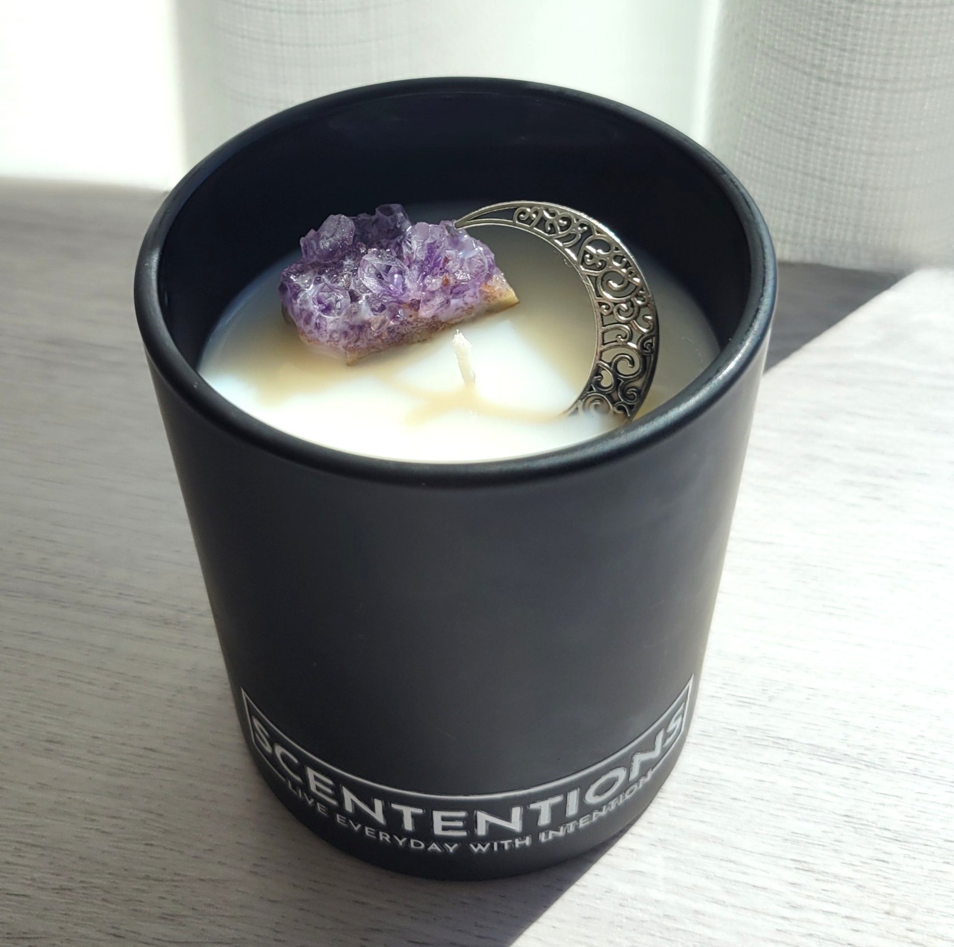 Moon + Bloom Moon Collection crystal coconut wax candle in matte black vessel with raw amethyst and silver moon charm in natural sunlight.