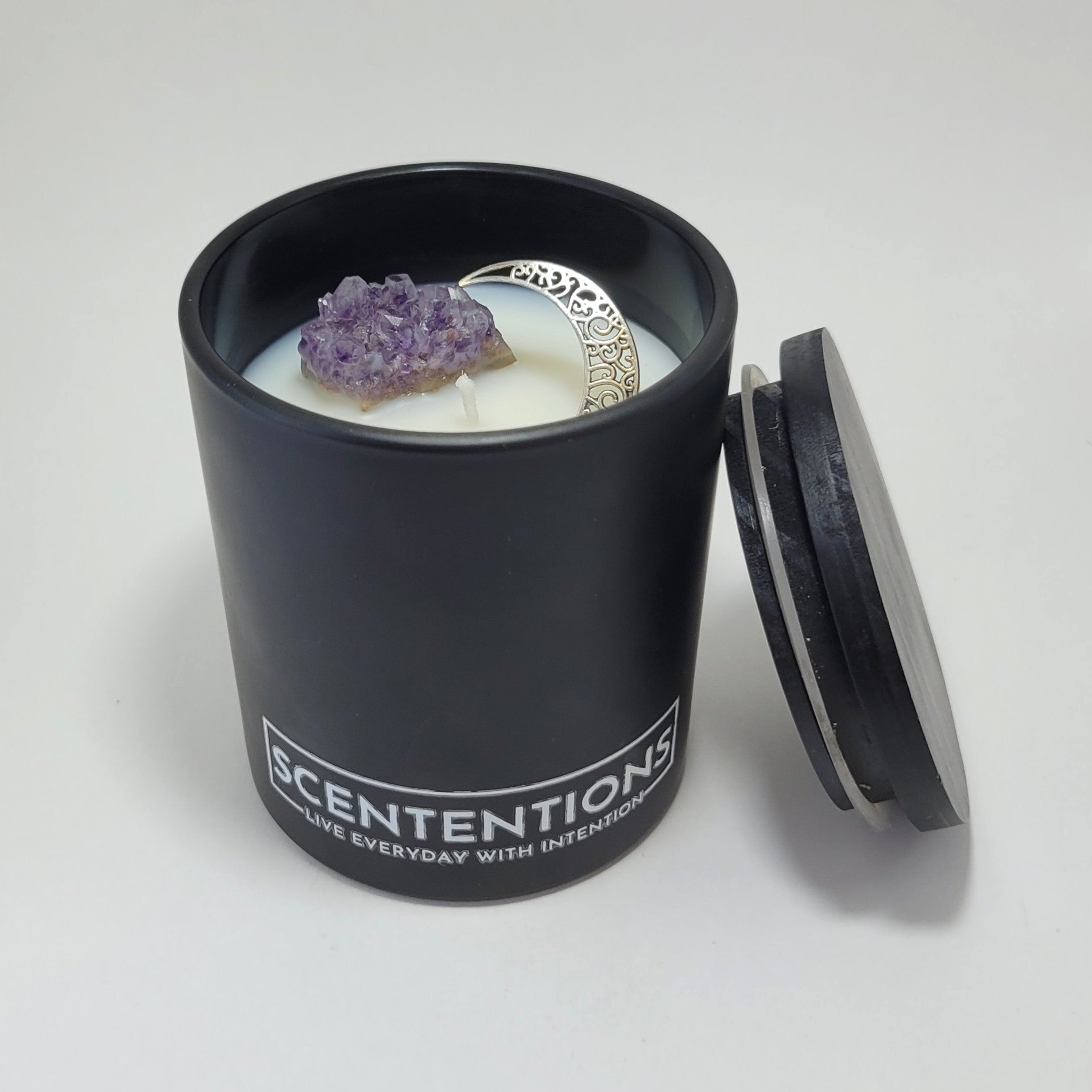 Moon + Bloom Moon Collection crystal coconut wax candle with raw amethyst, silver moon charm, and black wood lid beside jar.