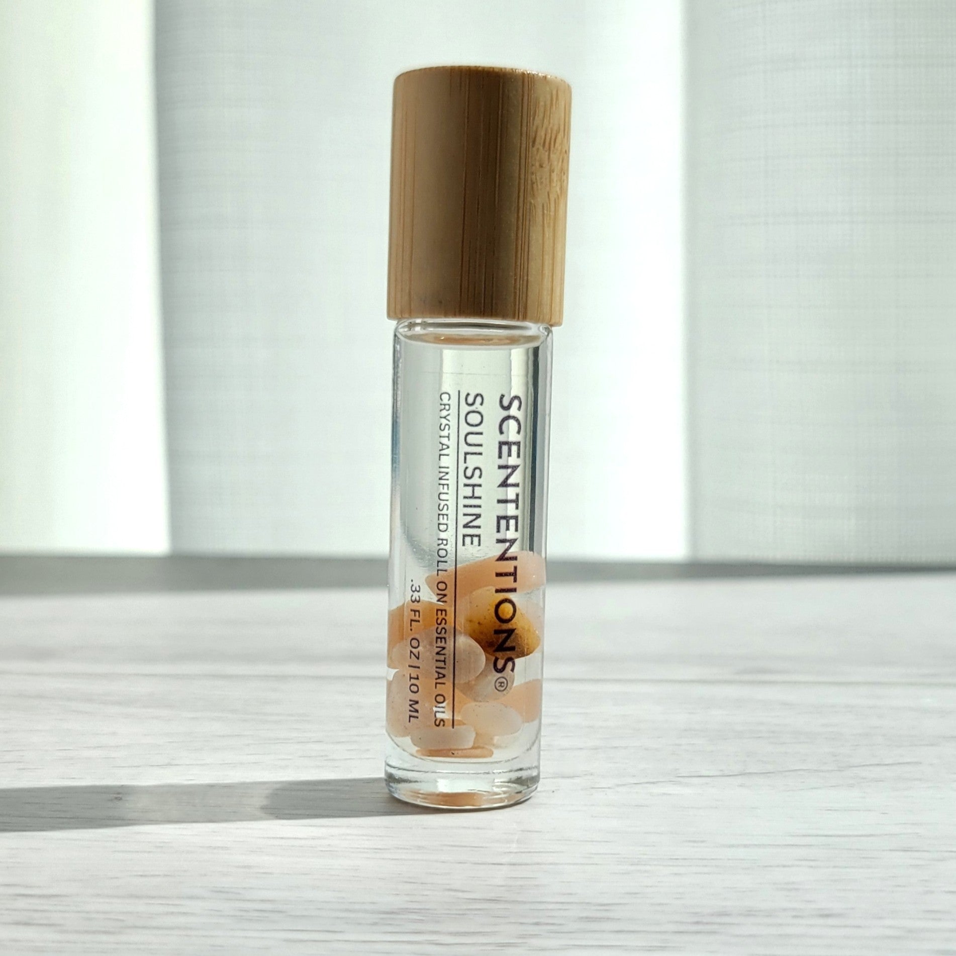 Crystal infused roll-on essential oil with Sunstone crystals and bamboo lid on white wood surface in natural light.